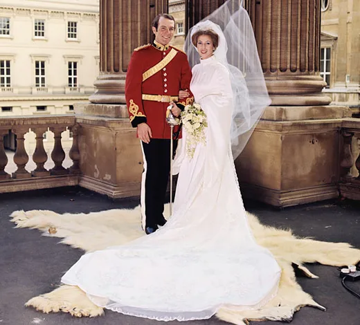 A bride in a flowing white gown and veil stands with a groom in red military uniform on a balcony, holding a bouquet.