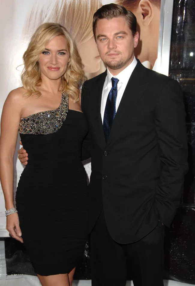 A man in a black suit with a woman in a black dress at a formal event, standing closely and smiling.