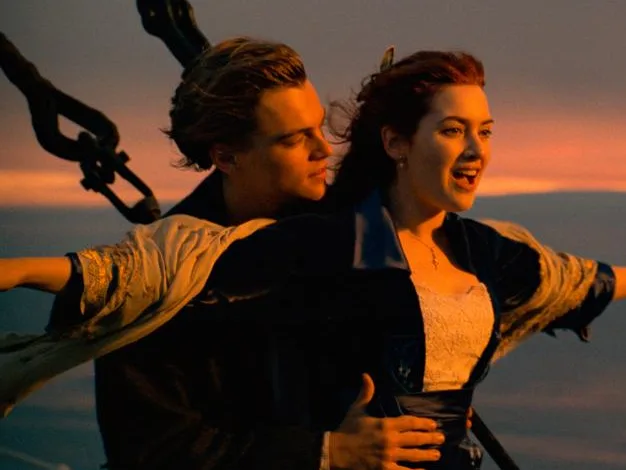 A couple reenacts the iconic Titanic scene on the ship's bow during a sunset.