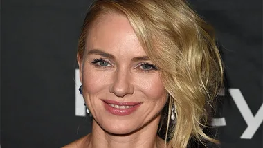 Naomi Watts smiling at an event, with a side-swept hairstyle and elegant earrings, against a dark backdrop.