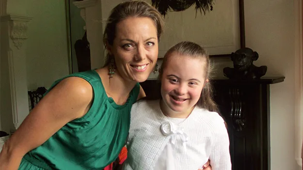 A woman in a green dress smiles with a young girl with Down syndrome in a white outfit, indoors near a fireplace.