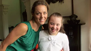 A woman in a green dress smiles with a young girl with Down syndrome in a white outfit, indoors near a fireplace.
