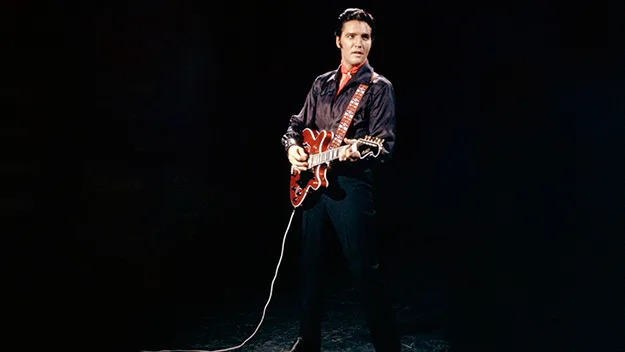 Elvis Presley performs on stage with a guitar, wearing a black outfit with a red shirt.