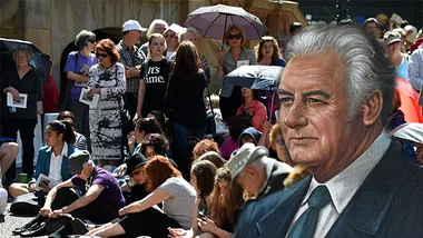 Crowd gathered under umbrellas for Gough Whitlam state memorial, with his portrait in the foreground.
