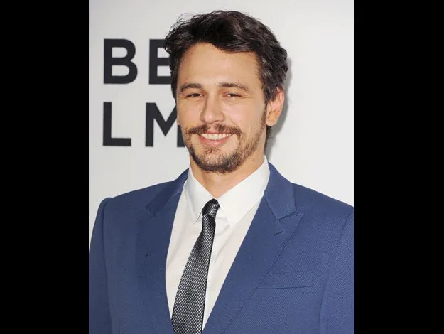 Man in a blue suit and tie smiles at an event with a white background.
