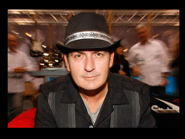 Man in a black hat and shirt sitting indoors with people blurred in the background.