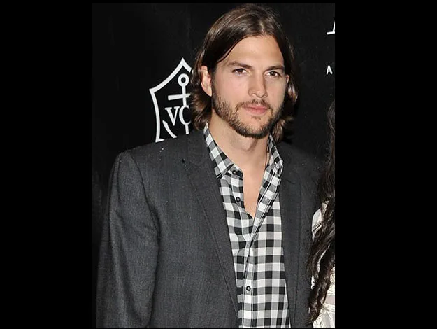 A man with shoulder-length hair and a beard, wearing a gray blazer and checkered shirt, stands in front of a dark backdrop.