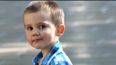 A young child with short hair wearing a blue checkered shirt, smiling slightly while looking at the camera.