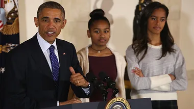 President speaking at a podium with two young women standing behind him, one in a burgundy top and the other in a gray sweater.