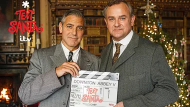 Two men in suits, one holding a "Downton Abbey V" clapperboard, with a Christmas tree in the background.