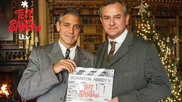 Two men in suits, one holding a "Downton Abbey V" clapperboard, with a Christmas tree in the background.