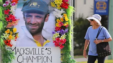 Portrait of Phillip Hughes with flowers and "Macksville's Champion" sign at a memorial, woman walking beside it.