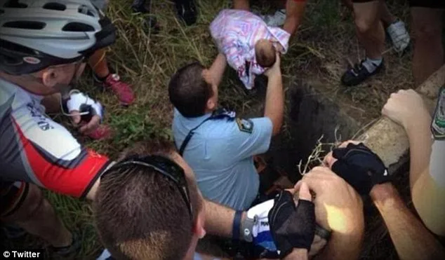 Rescue team pulling a wrapped infant from a drain surrounded by several cyclists assisting.