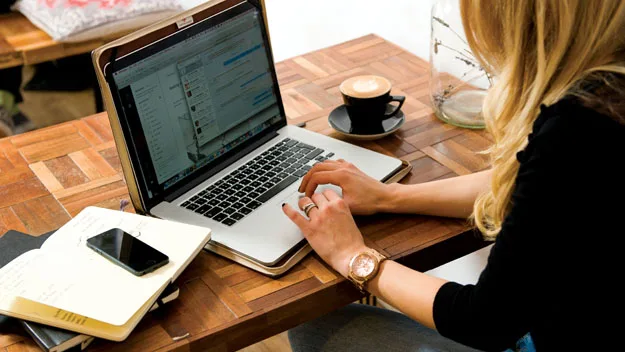 A woman working on a laptop in a cafe, with a cup of coffee and a notebook on the table.