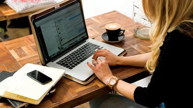 A woman working on a laptop in a cafe, with a cup of coffee and a notebook on the table.