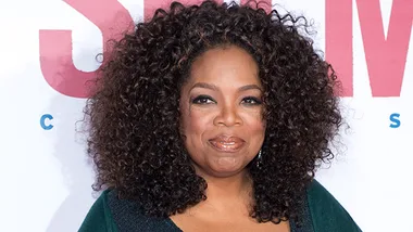 Oprah Winfrey smiling at a public event with a white background and red text partially visible behind her.