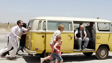 A family pushes a yellow van along a desert road, featuring a child in a red bandana from "Little Miss Sunshine."