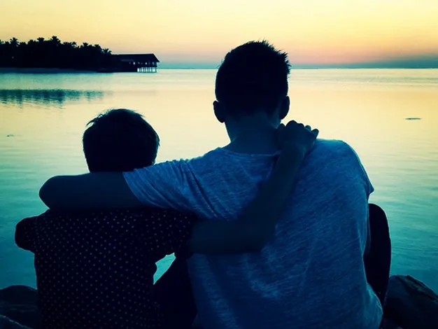 Two people sitting by a tranquil lake at sunset, with one person's arm around the other.