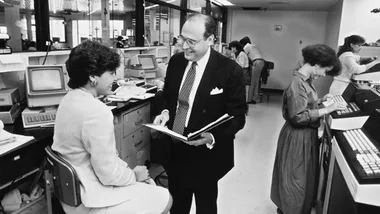 Businessman explaining something to a seated woman in an office, with people working at computers in the background.