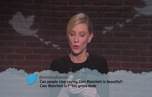 A woman reads a mean tweet about herself on a TV show, looking down at the paper against a backdrop with Twitter icons.