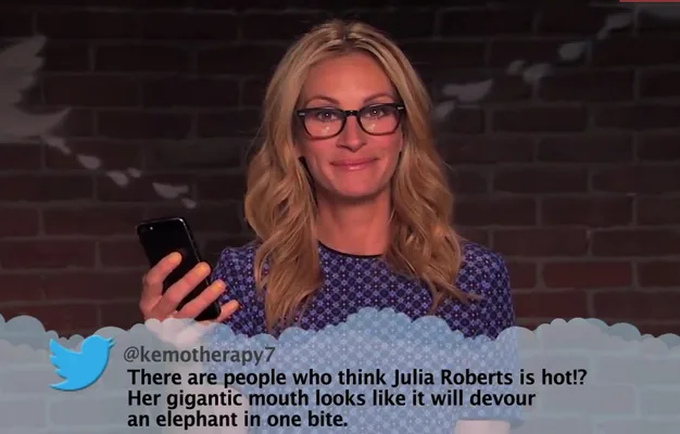 A woman in glasses reads a mean tweet about herself on a TV show, holding a phone with a brick wall backdrop.