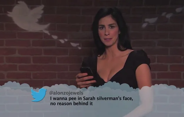 Comedian reading a mean tweet on a show set with a brick wall and blue Twitter bird graphic.