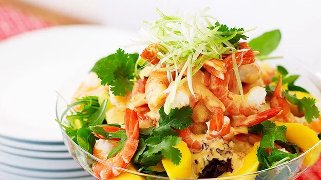 A glass bowl filled with shrimp salad, garnished with greens, cilantro, and lemon slices on a white plate background.