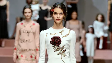 Model in white dress with "I love you mamma" embroidery, floral design, and rose hair accessory at Dolce & Gabbana runway.