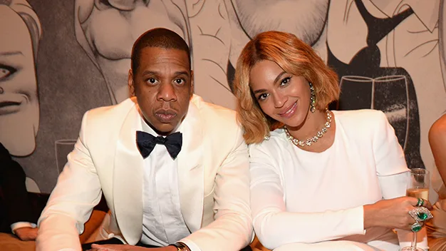 A couple in elegant white attire sits closely, the man in a bow tie and the woman holding a champagne glass, smiling warmly.