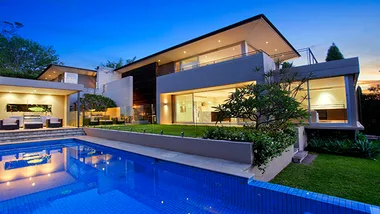 Modern two-story house with large windows, a pool, and outdoor seating, set against a twilight sky.