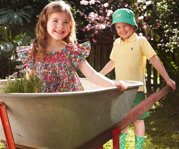 Two children in a garden, girl in floral dress in a wheelbarrow, boy in yellow shirt and green hat smiling beside her.