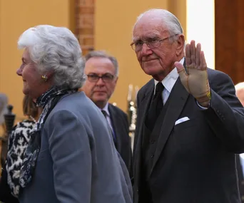 Elderly man in a suit waves, beside a woman in a grey outfit; both appear at a formal event.