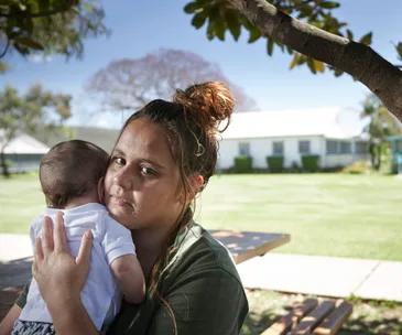 Woman in a green shirt holding a baby outdoors under a tree, with a house and trees in the background.