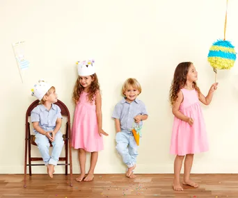 Children celebrate Easter with paper crowns and a piñata, two in pink dresses, one in blue shirt and crown, on a wooden floor.