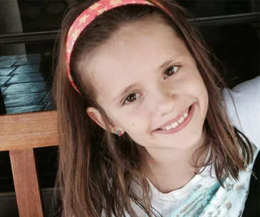 Young girl with long hair, wearing a headband and smiling while sitting on a wooden bench.