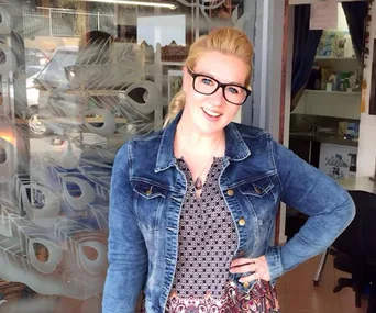 Woman smiling in a denim jacket and glasses, standing outside a shop with decorative glass.