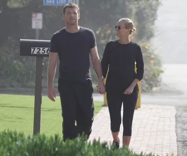A couple holding hands and walking outdoors on a sunny day, near a mailbox labeled 7256, with a road sign visible in the background.