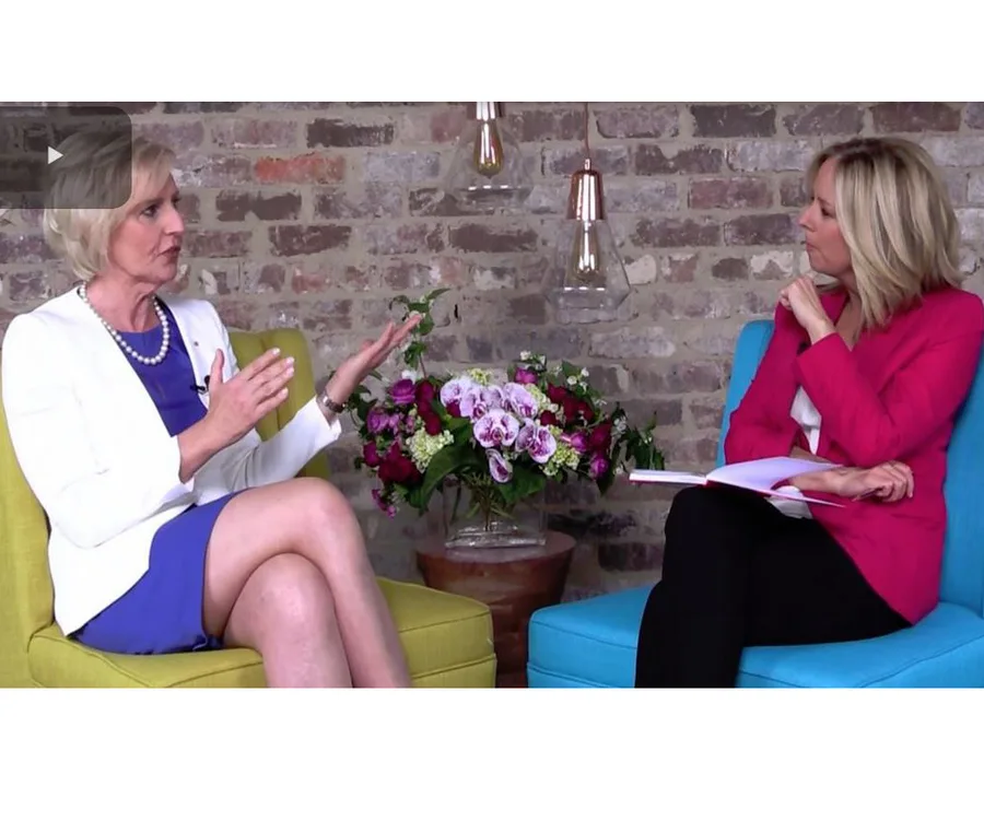 Two women are seated in a conversation setting, with a floral arrangement and brick wall in the background.