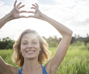 A woman smiling in a field, making a heart shape with her hands above her head.