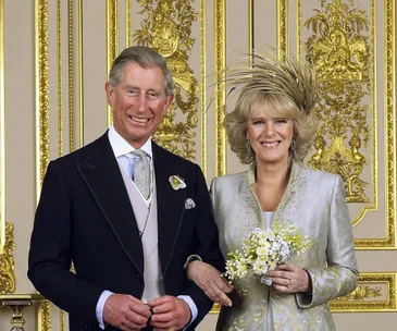 Prince Charles and Camilla standing in formal attire, smiling, with ornate gold decor in the background.