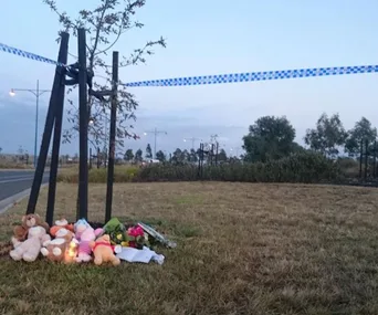 A makeshift memorial with teddy bears, flowers, and a candle by a roadside, cordoned off with police tape.