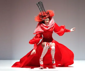 Model in dramatic red outfit and tall headpiece stumbles on runway, showcasing avant-garde fashion design.
