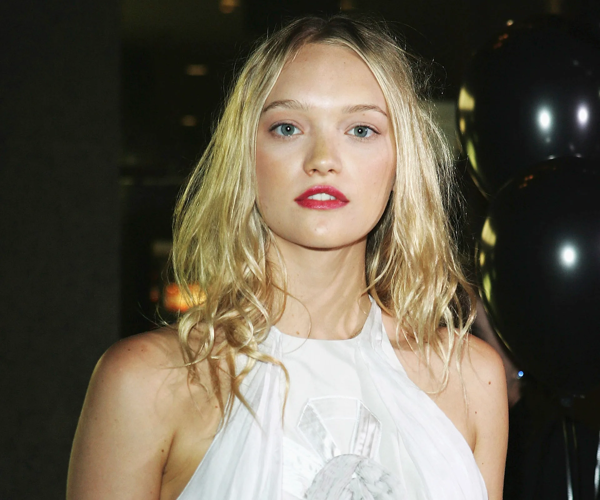Blonde woman in a white dress with red lipstick standing indoors near black balloons.