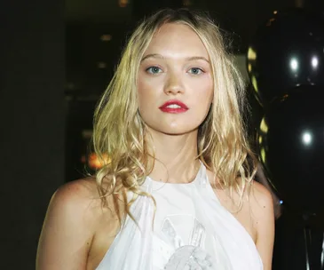 Blonde woman in a white dress with red lipstick standing indoors near black balloons.