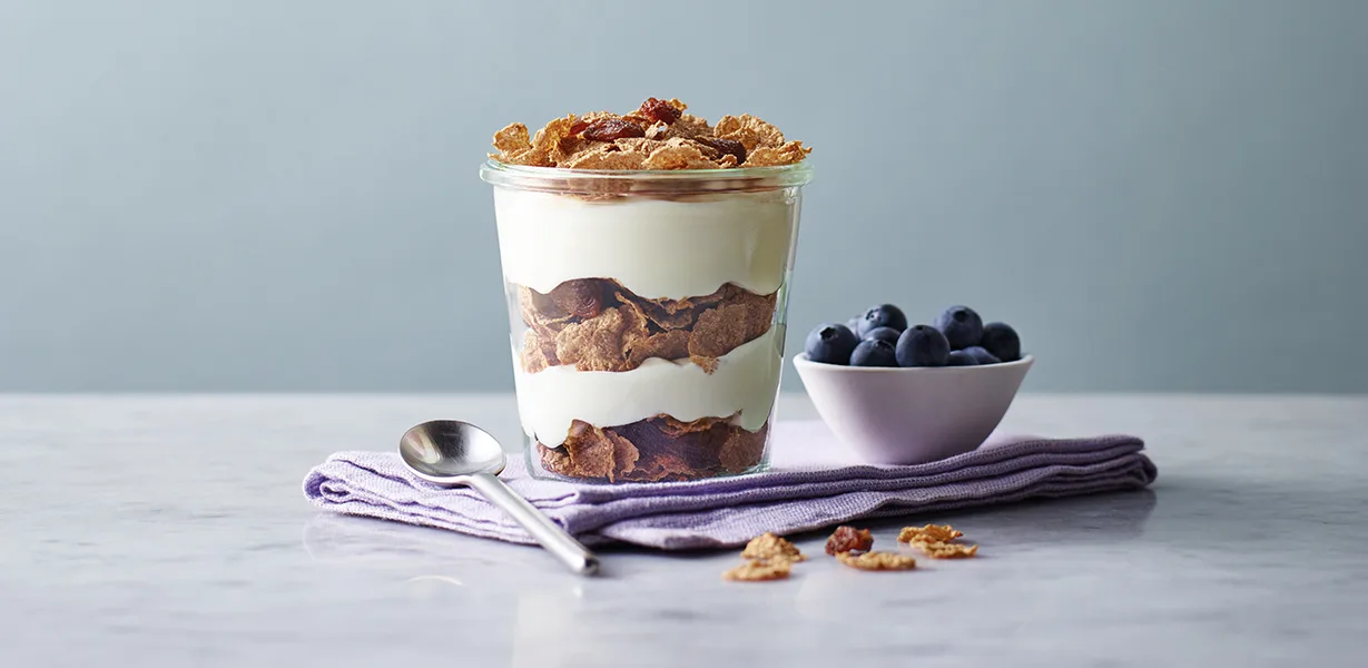Glass jar with layered yogurt and cereal, spoon beside it on a napkin, bowl of blueberries nearby.
