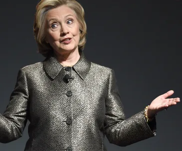 A woman in a patterned jacket gestures with both hands while speaking.