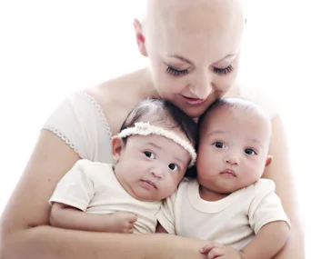 A mother with no hair gently holds her twin babies, one with a white headband, close to her in a loving embrace.