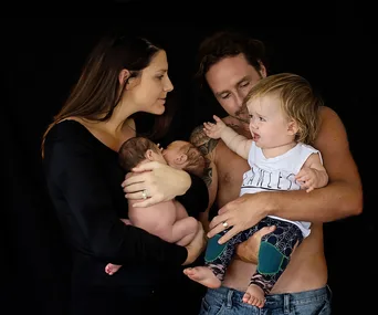 Family photo with two adults holding a toddler and two newborn twins, against a black background.