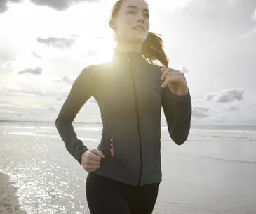 A woman jogging on a beach wearing a gray athletic jacket, with sunlight shining behind her.