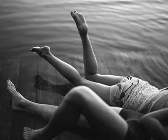 A black and white photo of two people lounging on a dock, with legs playfully extended over the water.